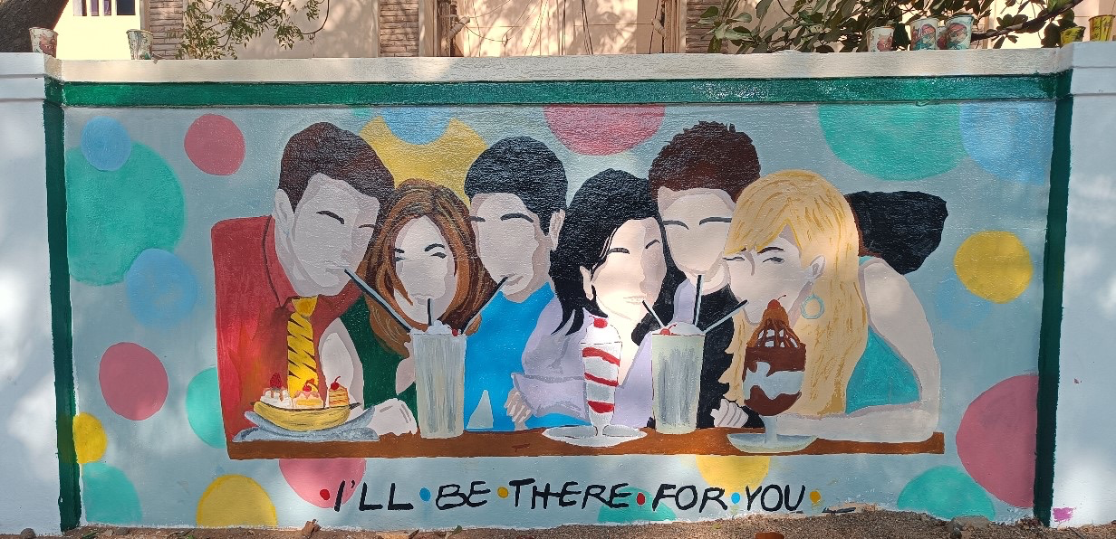 A mural painting on an outdoor wall features six friends sitting together at a diner table, sharing milkshakes and desserts, with three people sharing a single drink. The painting is inspired by the TV show Friends, depicting stylized versions of the main characters with minimal facial details. The background has colorful polka dots, and at the bottom, the phrase "I'LL BE THERE FOR YOU" is written, referencing the show's theme song. 