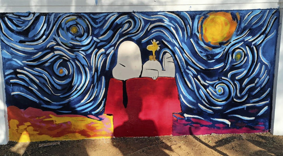 A mural painting on an outdoor wall depicts Snoopy lying on his back on top of his red doghouse, while Woodstock is sitting on his belly. 
Woodstock is gazing at a night sky inspired by Van Gogh's "The Starry Night." The swirling blue sky features a large yellow sun, while the lower part of the mural has warm tones of orange, pink, and yellow, resembling a landscape.