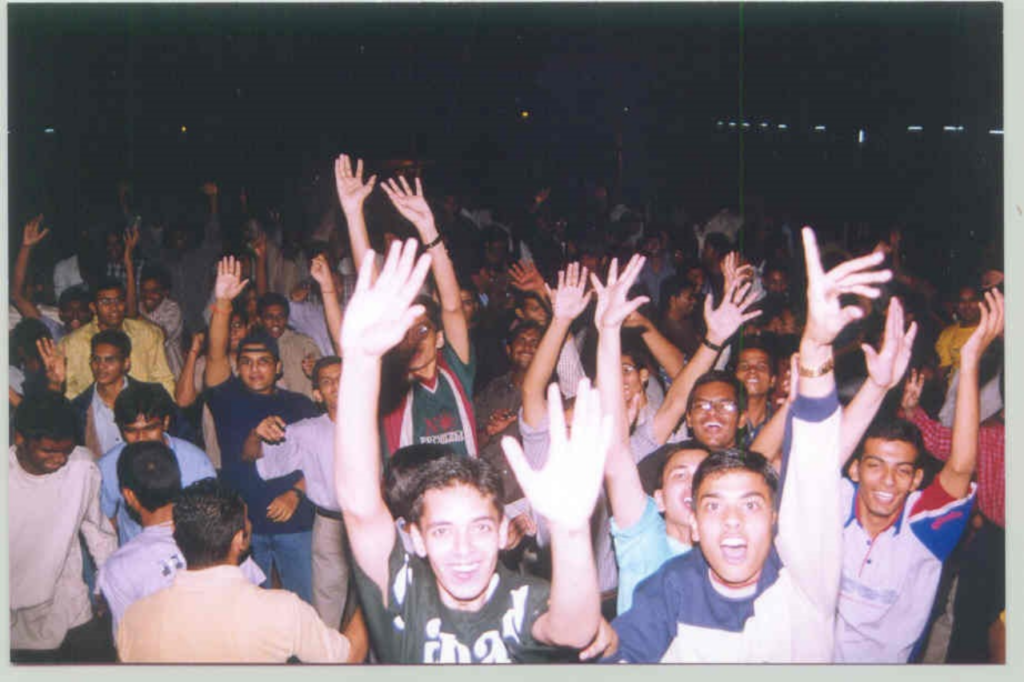 A crowd of teenagers look into the glare of the camera with their hands raised. They seem to be celebrating, just like the rest of the crowd behind them