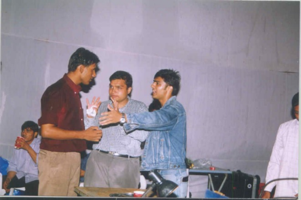 Three people appear to be arguing, all looking at each other. The paunchy young man in the centre appears to be placating the other two men on either sides of him.