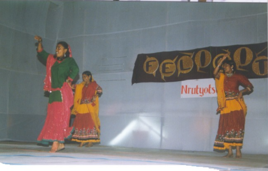 Three girls, dressed in traditional clothes, are dancing in formation. From the perspective of someone photographing from the level of the stage, there is one person in front, while two others are in the second row.