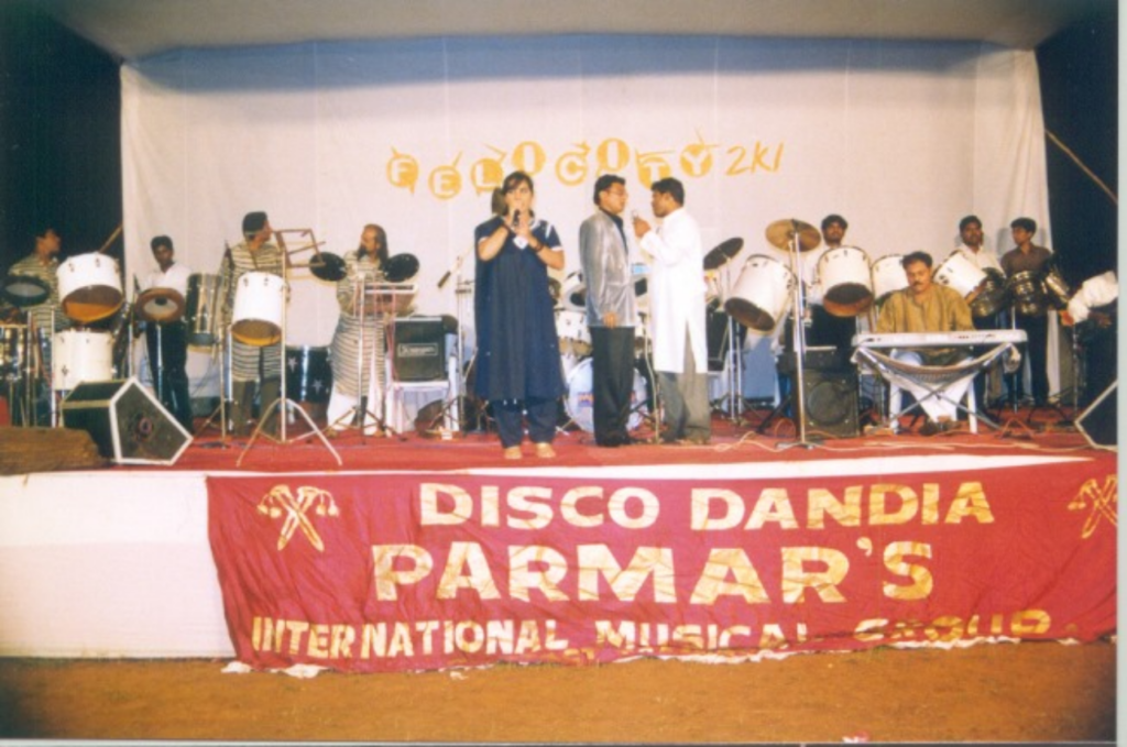 A horde of instruments crowd the stage that is being pictured. In the centre, in blue, stands a women holding the mic, presumably to sing. Below the level of the stage, a red banner is draped, reading "Disco Dandiya / Parmar's / International Music Group" in golden lettering. Behind the band is the logo of Felicity 2k1 in golden letters on a white background