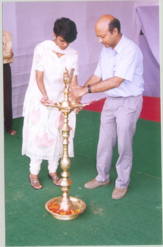 A bald middle-aged man, dressed in a shirt and pant, is lighting a traditional Indian lamp with the help of a young lady in a white salwaar kameez