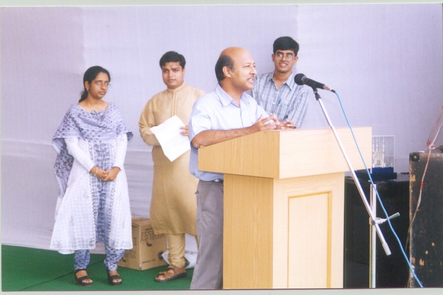 A balding, middle-aged man adreeses the (invisible) crowd from a podium. Onlooking him in the background are 3 people.