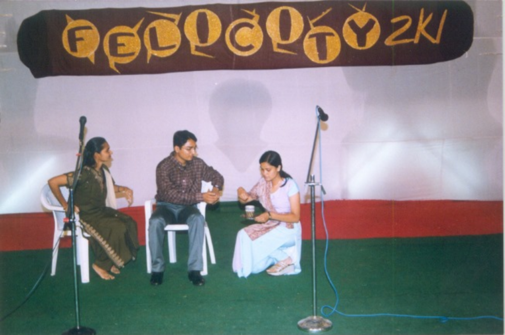 The picture is of a stage. Towards the left (of the viewer), a young gentleman and lady seated on chairs. Both are looking at another lady, who is kneeling down and holding a tray with a glass and some food. In the background, Felicity 2k1 is written with a fancy font.