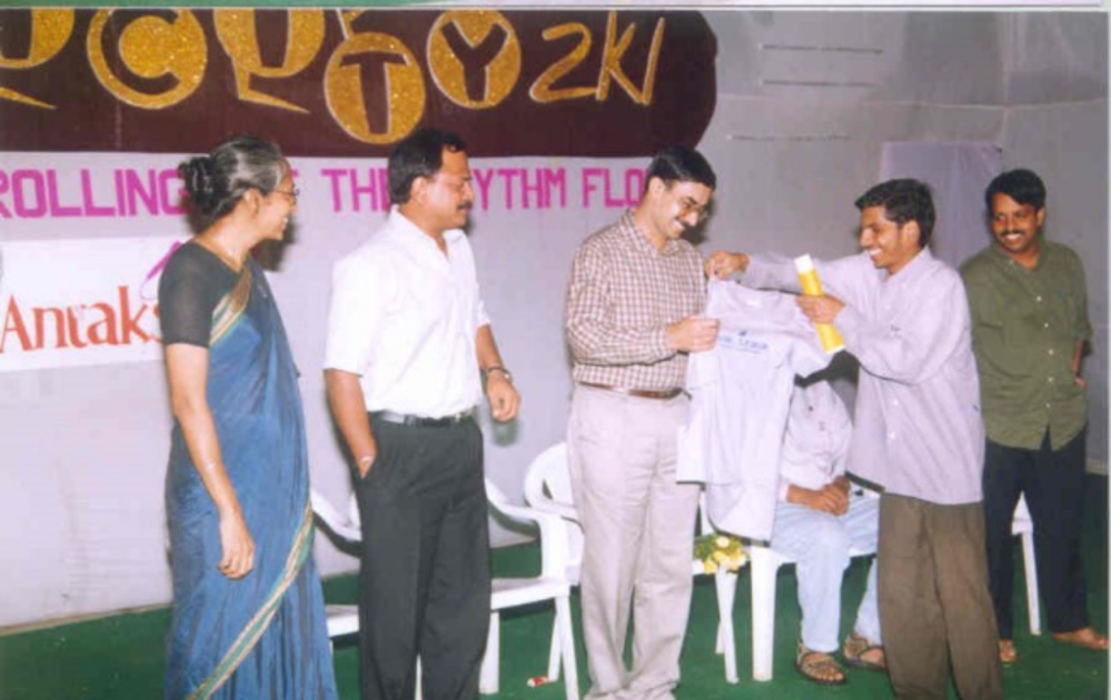 Five people are standing in a line on the stage, where a sixth person is handing out a T-shirt to the person in the centre. Others look on joyfully.